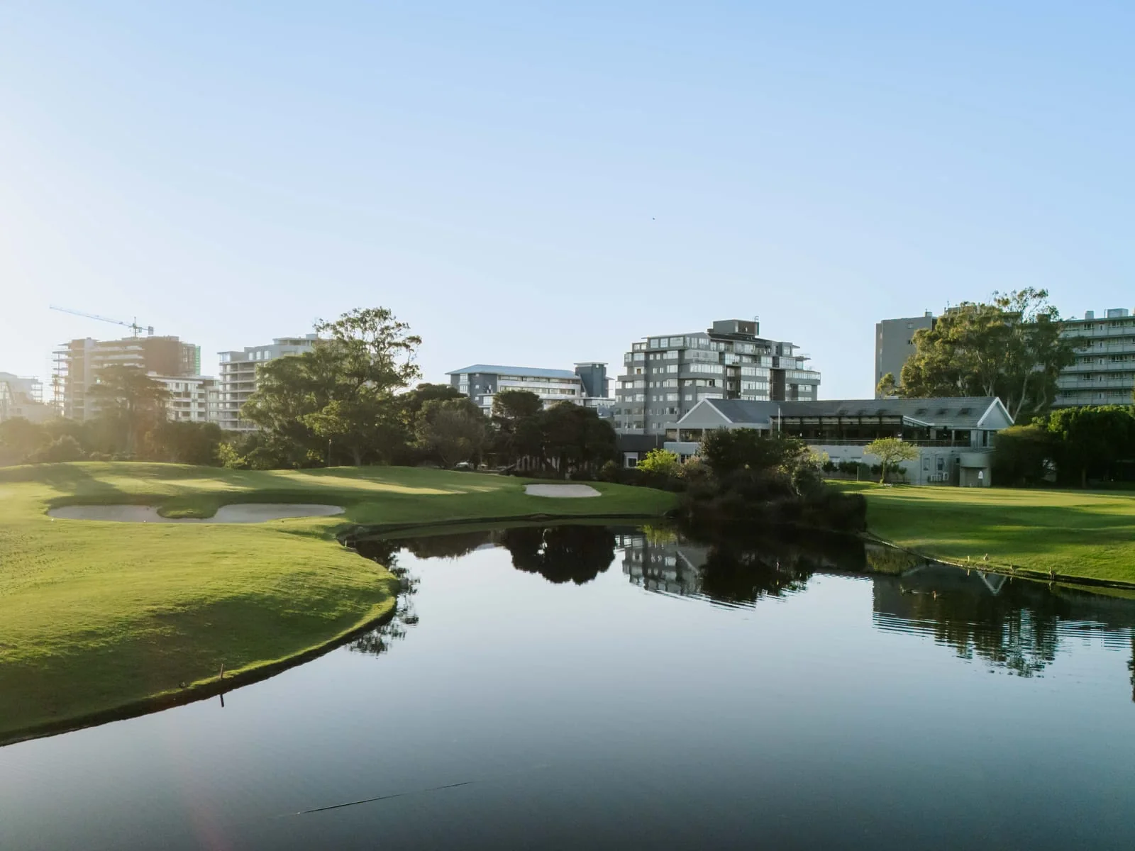 A still pond beside a green and its bunkers, with apartments beyond